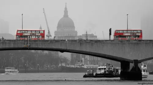 The photograph is an urban landscape taken in London, United Kingdom, during the early afternoon in winter. The image prominently features two iconic red London buses crossing a city bridge, set against the architectural backdrop of St Paul's Cathedral. The bridge, typical of London architecture, spans the River Thames and is surrounded by various riverboats and barges on the water. The misty, overcast atmosphere and bare trees in the scene suggest a cold winter's day. St Paul's Cathedral, a famous landmark, rises above the skyline and is visible through the haze, adding to the distinct London cityscape. The photograph captures the energy of city life and urban transportation in London, highlighting key elements such as the bridge, buses, and the historic cathedral in the heart of the United Kingdom's capital.